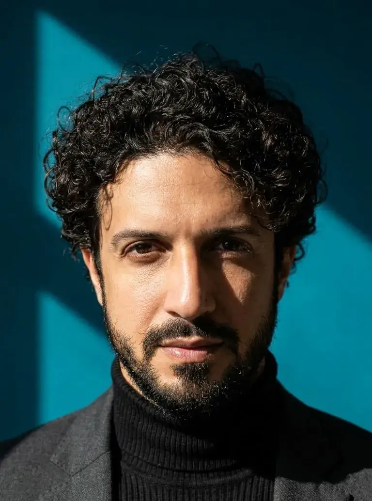 Retrato em close-up de Yan Almeida de Carvalho (Yan Carvalho), criador da Ad Quem, com cabelo cacheado escuro, barba curta e terno escuro sobre blusa de gola alta, posando contra um fundo azul esverdeado iluminado por luz natural.
