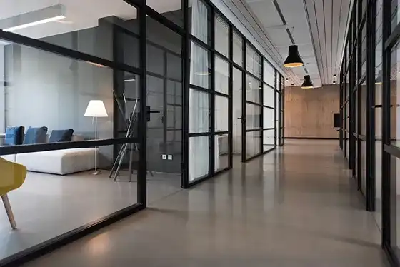 Corredor moderno de escritório com divisórias de vidro e estruturas metálicas pretas, piso cinza polido e salas com sofás e luminárias pendentes.